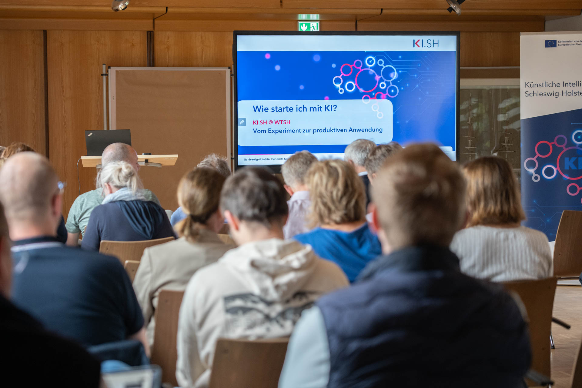 viele Menschen sitzen in Stuhlreihen vor einer KI.SH-Beamerleinwand